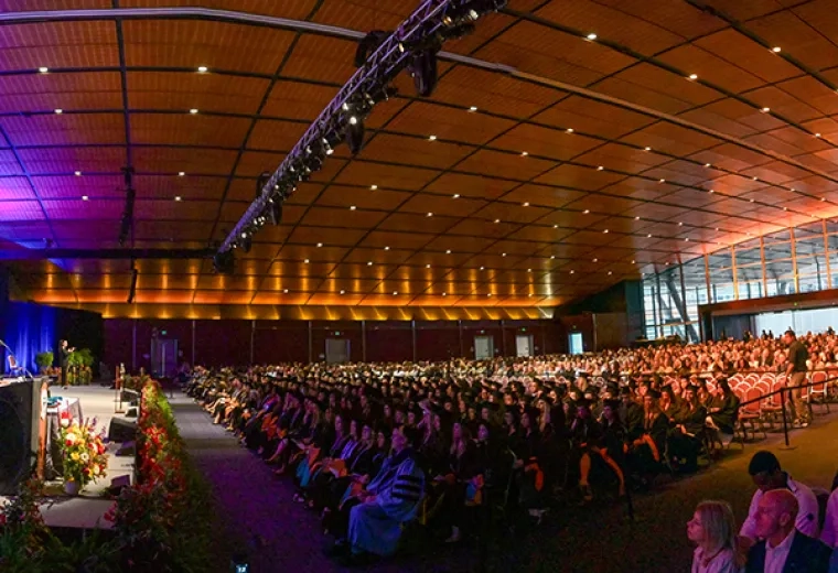 A large room with people dressed for commencement and an audience behind them