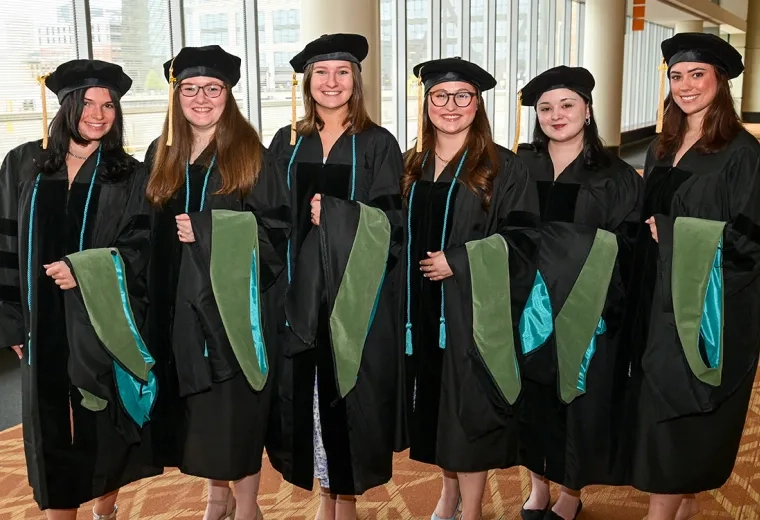 Six women wearing graduation gowns and holding academic hoods pose for the camera at commencement