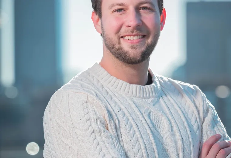 Headshot of Ian Karby standing with his arms crossed against a city skyline background wearing a white sweater and a watch.