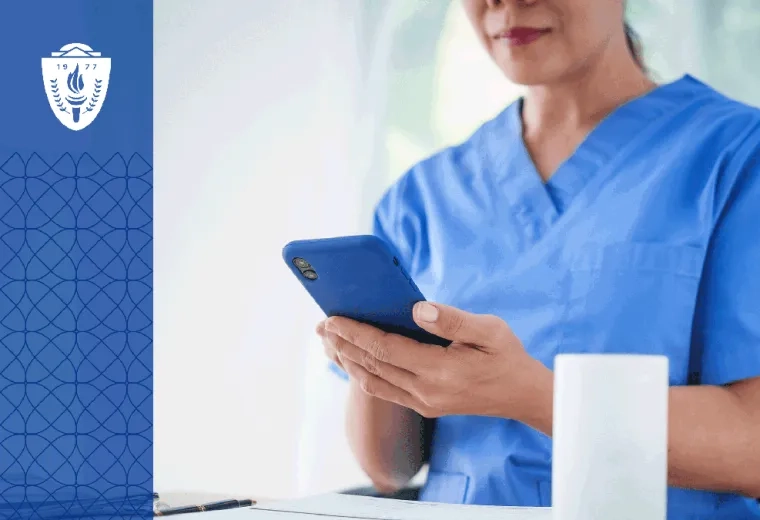 Woman in blue scrubs sitting a desk looking at her phone