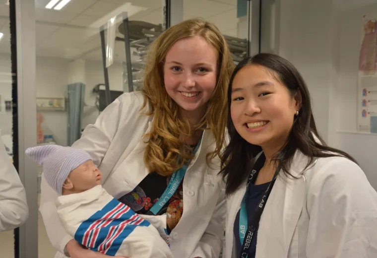 Two female students in white coats pose with a newborn baby manikin