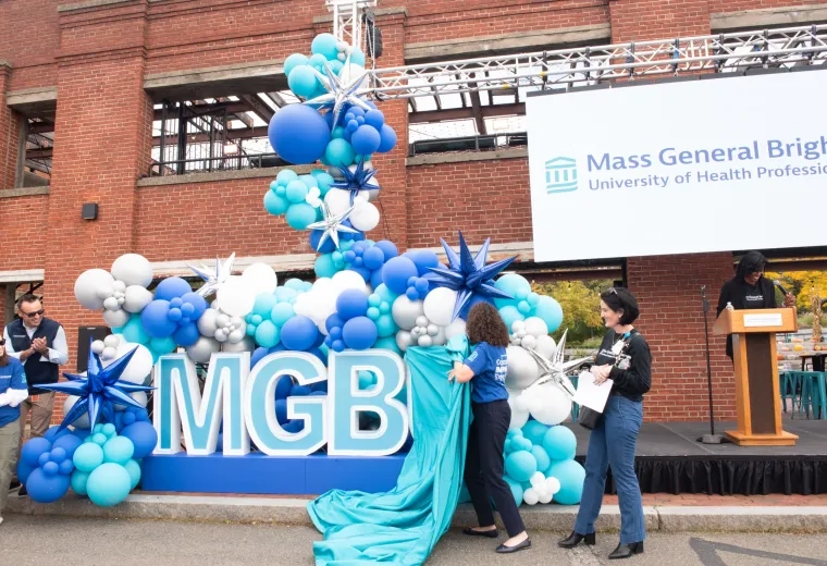 Two women pulling a blue sheet off of letters that say MGBU
