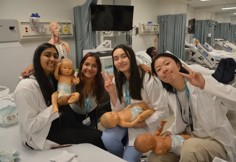 Four female students in white coats pose in a emergency room while holding infant manikins