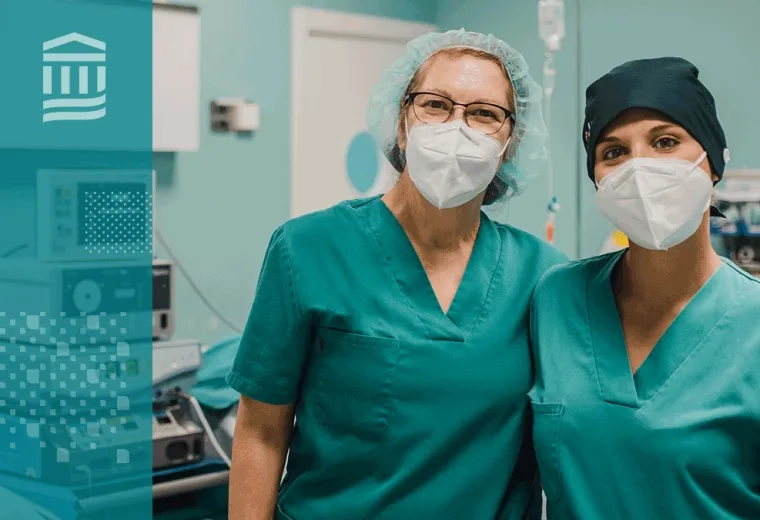 two women with masks and green scrubs stand arm and arm