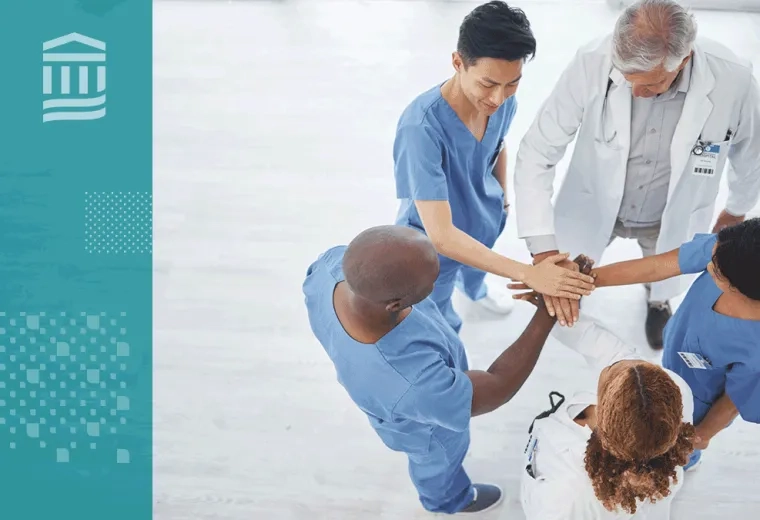 Members of a healthcare team stand in a circle with their hands stacked together in a show of teamwork.
