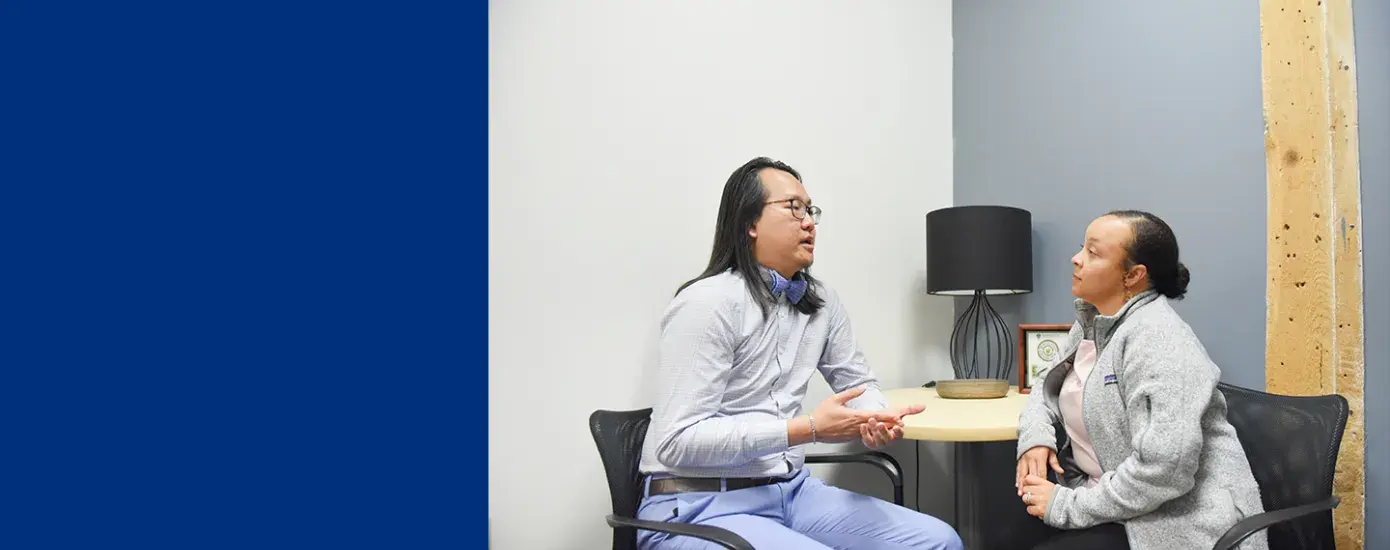 man of Asian descent sits and speaks with a woman with curly hair and brown skin