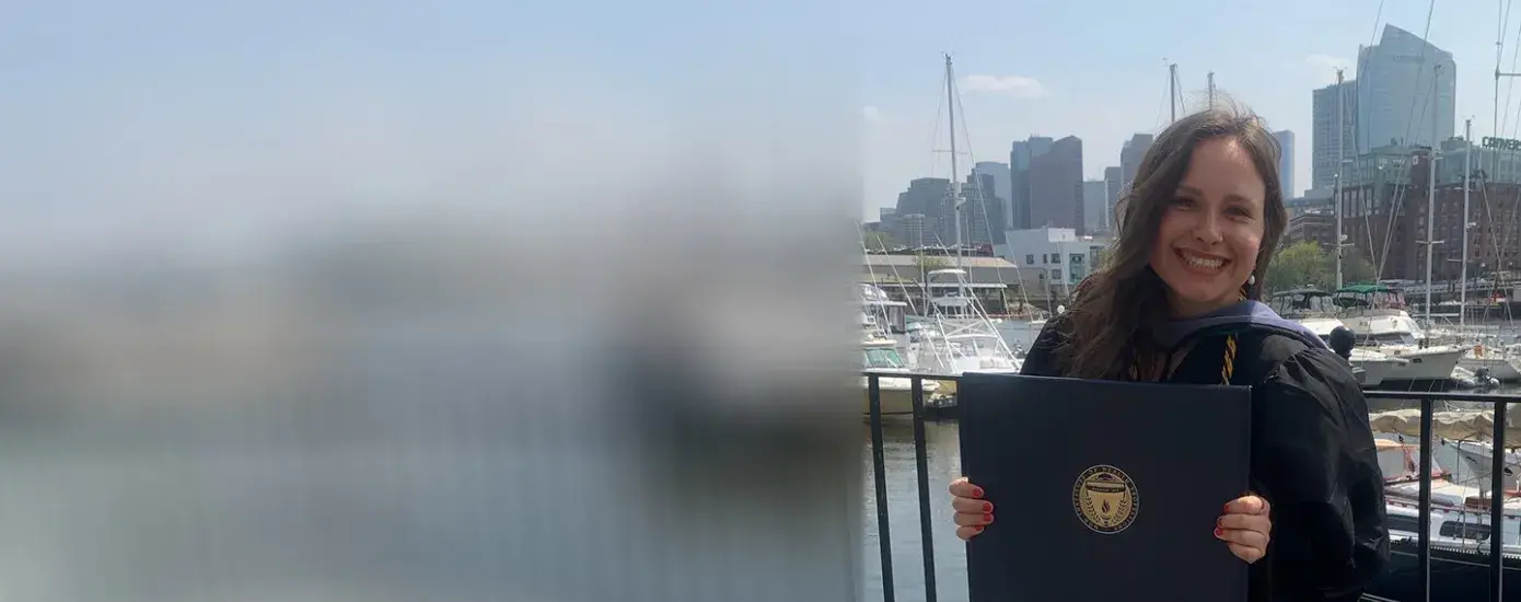 A woman wearing a graduation gown holds a diploma in front of a background of sailboats