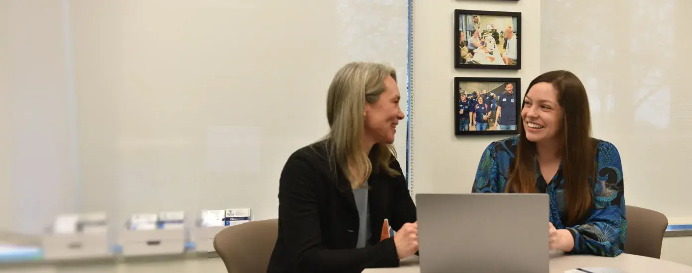 Two women sit at a desk talking with a computer between them