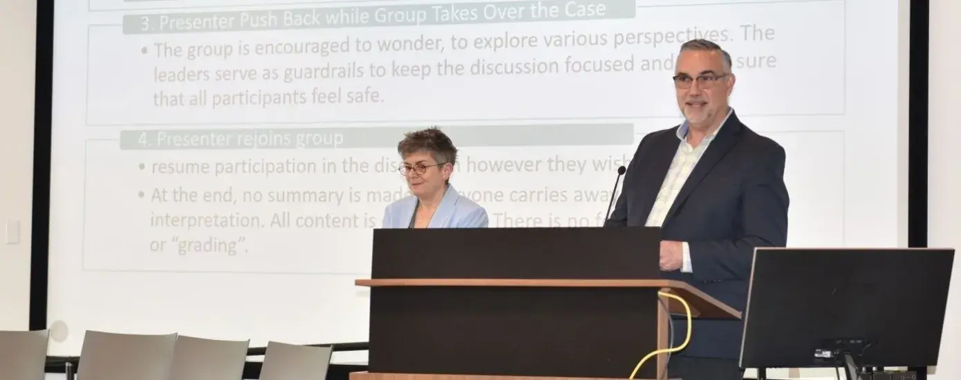 A man stands at a podium with a woman standing next to him and empty seats to his right