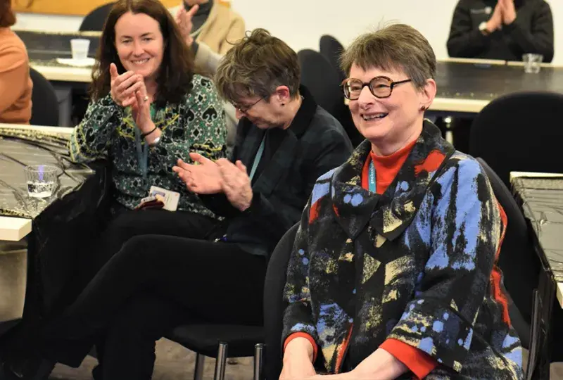 woman with glasses seated at a table smiles broadly as people clap around her