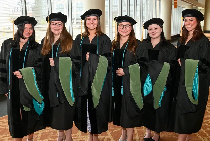 Six women wearing graduation gowns and holding academic hoods pose for the camera at commencement