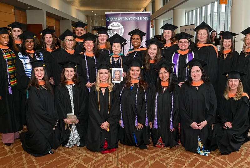 23 people wearing graduation gowns stand in three rows for a commencement photo