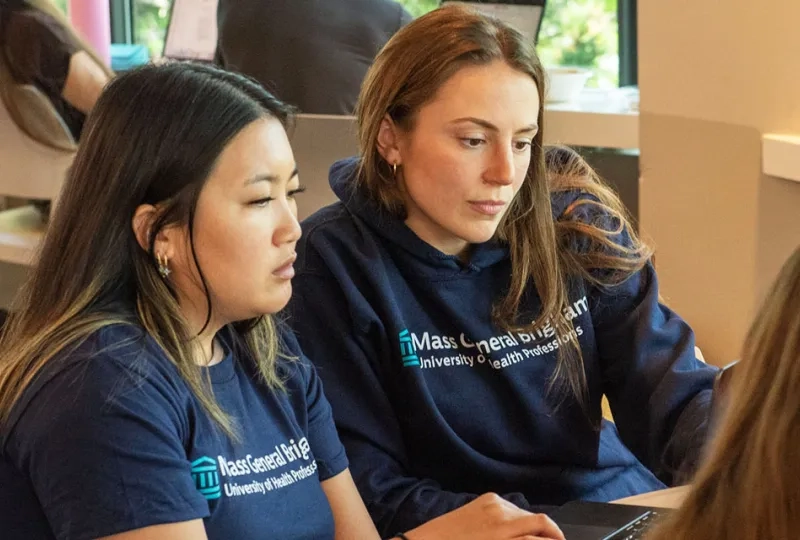 Two Mass General Brigham University students work during class wearing T-shirts with the university's name