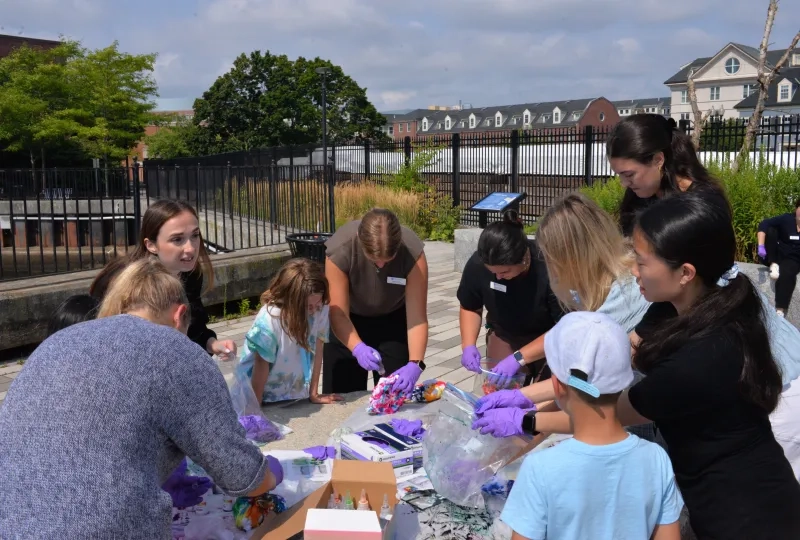 Kids and adults wear purple gloves and stand around a table dying shirts