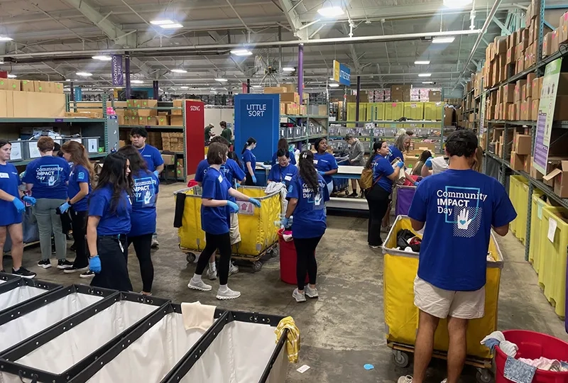 Volunteers in blue shirts that read "Community Impact Day" sort clothes and supplies into large yellow bins in a warehouse