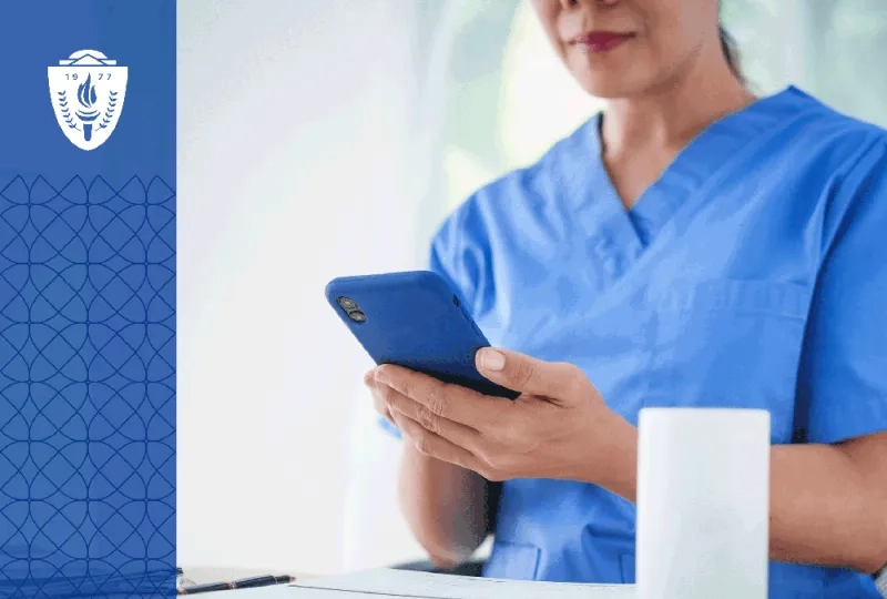 Woman in blue scrubs sitting a desk looking at her phone