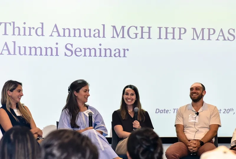 A panel of alumni sits on a stage holding microphones with the words Third Annual MGH IHP MPAS Alumni Seminar beind them