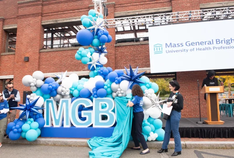 Two women pulling a blue sheet off of letters that say MGBU