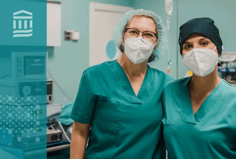 two women with masks and green scrubs stand arm and arm