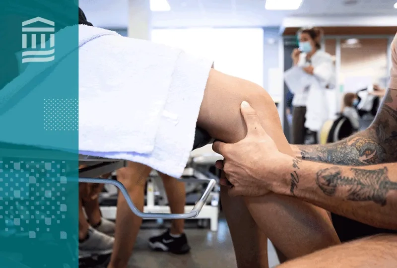 a person sits in front of another who is on a medical table, the focus are the person's hands around the other's knee