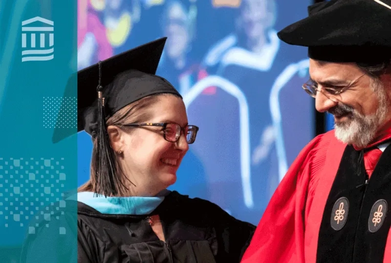 professor in graduaiton regalia (cap and red gown) smiles at a woman graduating who is in blue regalia