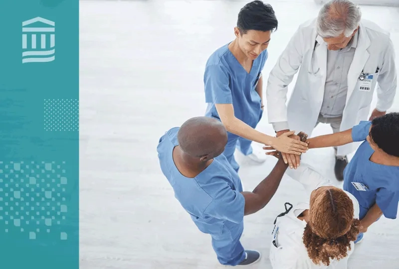 Members of a healthcare team stand in a circle with their hands stacked together in a show of teamwork.