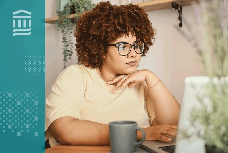 Woman wearing a yellow shirt sitting at a desk working on a laptop next to a coffee mug