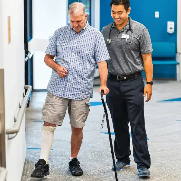 student helps older man with a cane to walk down a hall