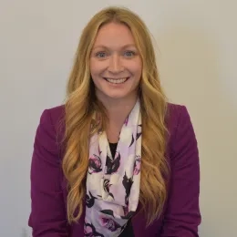 Headshot of woman with long blonde hair standing in front of solid white background wearing a light purple scarf and dark purple jacket