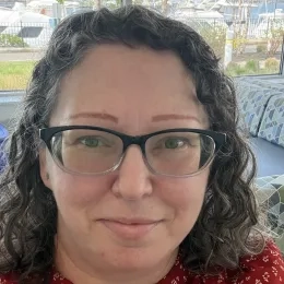Head shot of Woman with curly dark hair wearing glasses and a red shirt in front of a waterfront background.