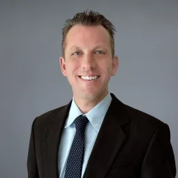 Headshot of Jason Richter standing against a solid gray background, wearing a blue collared shirt, a black blazer and, and blue necktie with white dots
