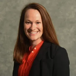 Headshot of Adrian Wright-FitzGerald with long dark blond hair, wearing an orange shirt and a black jacket against a gray background