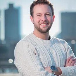 Headshot of Ian Karby standing with his arms crossed against a city skyline background wearing a white sweater and a watch.