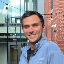 Headshot of Anthony Sposito wearing a blue MGH sweatshirt in front of a brick and glass building in the background