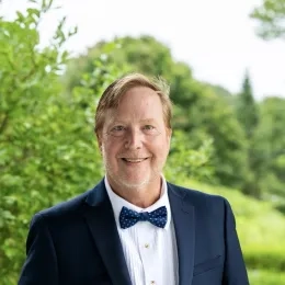 Headshot of Scott Goodspeed wearing a white button down shirt, dark blue blazer, and blue bowtie. He is standing in front of a background of green grass and trees