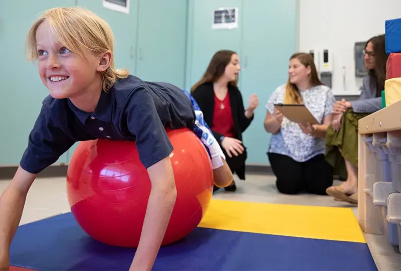 A blond child balances on his stomach on a big red ball while women talk in the background