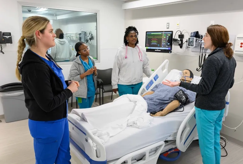 nursing students stand around a manikin in a hospital bed and listen to a teacher speak