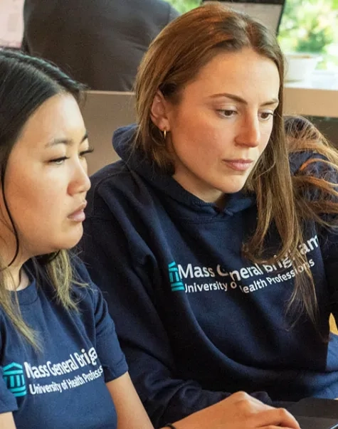 Two Mass General Brigham University students work during class wearing T-shirts with the university's name
